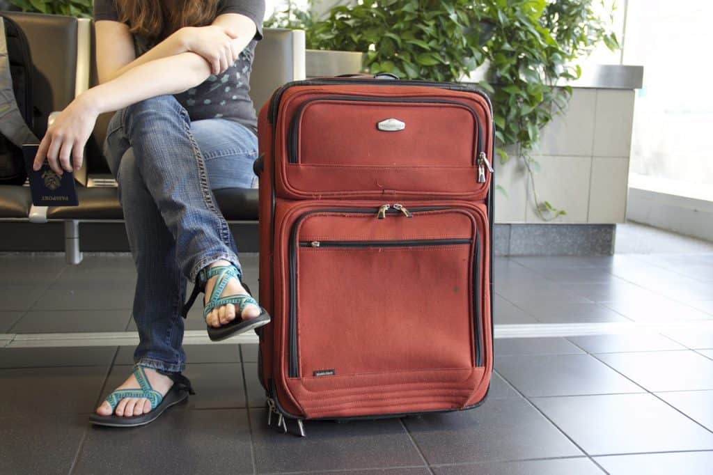 A person wearing jeans and sandals waits in the airport with their large red suitcase.
